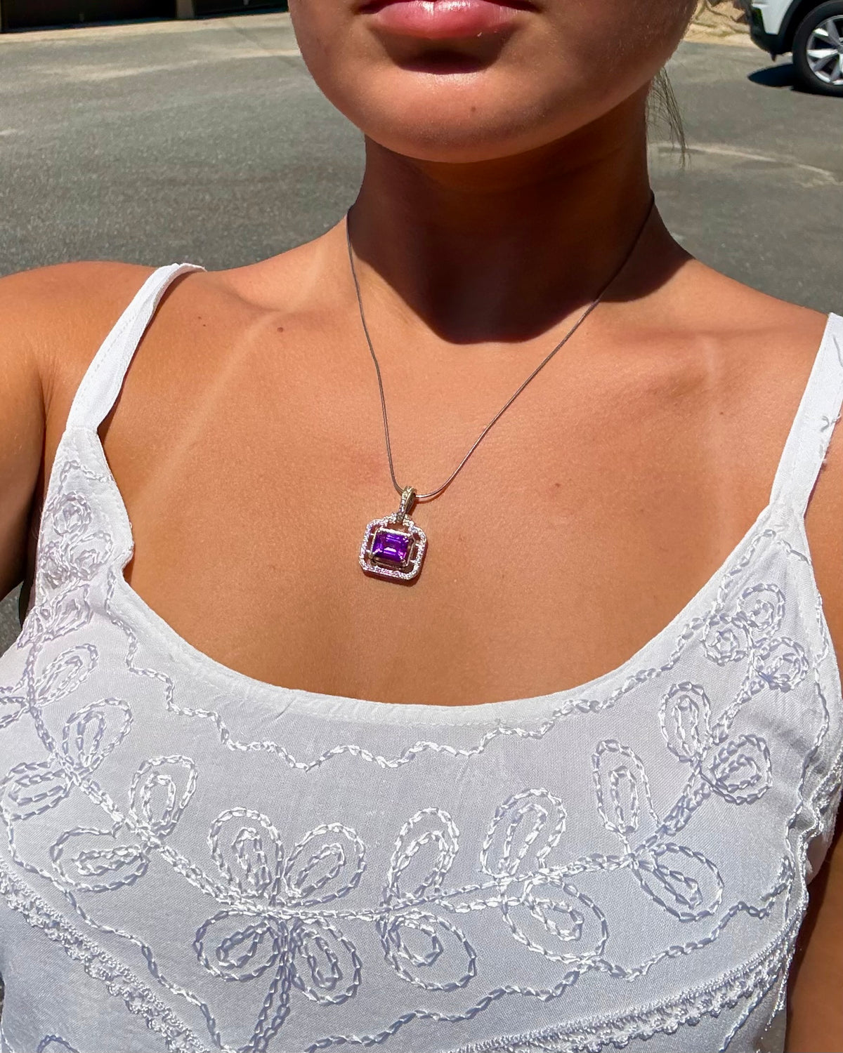 A close-up lifestyle shot of a woman wearing a silver-toned necklace with a square purple gemstone pendant, styled with a white embroidered tank top.